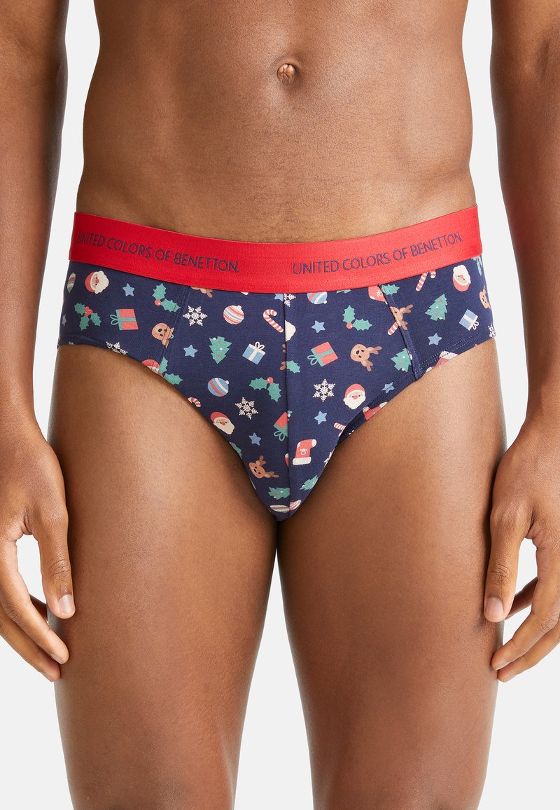 Uomo che indossa slip blu navy con stampe a tema natalizio e una vita rossa con l'etichetta "United Colors of Benetton."