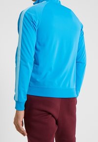 Blaues Sportjacke mit hohem Kragen, glatter Textur und hellblauen Streifen an den Ärmeln. Kombiniert mit dunkelroten Hosen.
