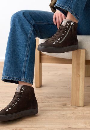 Personne assise sur une chaise en bois portant un jean bleu et des baskets montantes marron foncé avec des fermetures éclair latérales sur un sol en bois clair.