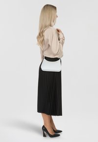 Borsa a tracolla bianca con superficie liscia e forma curvilinea, indossata con una blusa beige e una gonna plissettata nera, completata da tacchi neri.