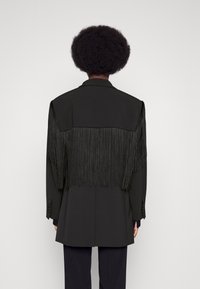 Veste noire avec un long détail de franges sur les épaules, texture lisse et coupe structurée. Design ajusté avec poches latérales.
