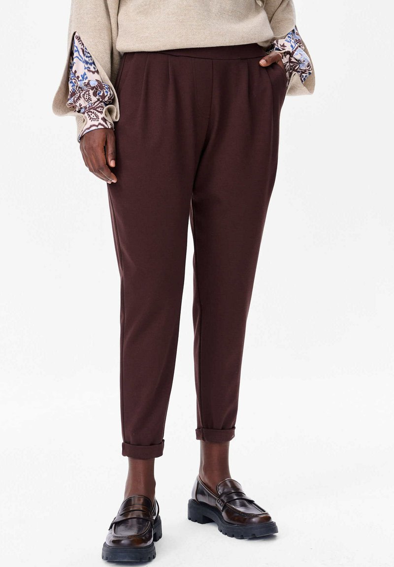 Brown, tailored trousers with a tapered fit, featuring a smooth texture and rolled cuffs. Paired with dark loafers and a beige top.