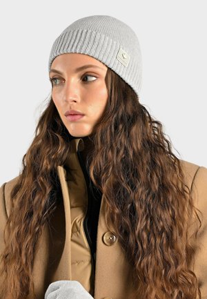 Bonnet côtelé gris en tissu tricoté doux, avec un petit logo. La personne porte des cheveux longs et ondulés et est habillée d'un manteau beige.