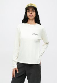 Langarmshirt in roomkleur, gemaakt van zachte stof, met zwarte "OBEY"-tekst en steraccents op de linkerborst. Gecombineerd met een donkere broek en een gele pet.