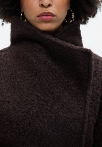 Abrigo marrón texturizado con un alto cuello, que presenta un acabado suave y peludo con costuras mínimas. Accesorio: grandes pendientes de aro dorados.