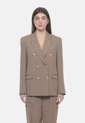 Donna che indossa un blazer doppiopetto color sabbia con bottoni dorati e pantaloni abbinati, in piedi davanti a uno sfondo bianco uniforme.