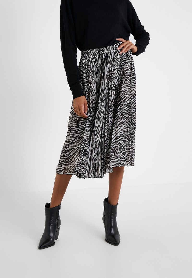 Femme portant un haut à manches longues noir, une jupe midi plissée à motifs noir et blanc, et des bottines noires à talons hauts.