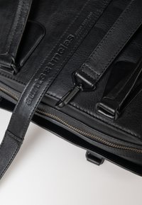 Schwarze Ledertasche mit strukturiertem Finish, verstellbaren Trägern, Reißverschluss und geprägtem Markenlogo entlang des Trägers.