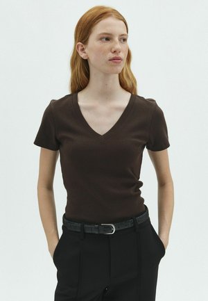 Jeune femme aux cheveux roux portant un t-shirt marron foncé à col en V et manches courtes, et un pantalon noir avec une ceinture noire, les mains dans les poches, regardant sur le côté.
