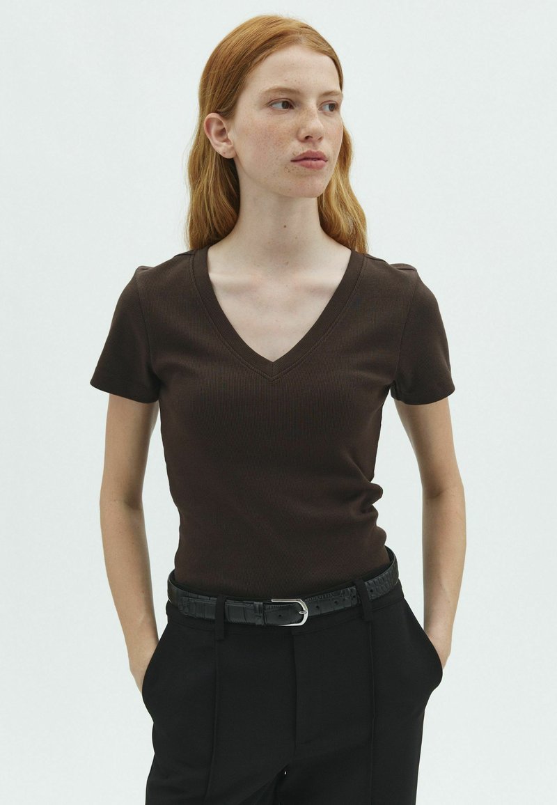 Jeune femme aux cheveux roux portant un t-shirt marron foncé à col en V et manches courtes, et un pantalon noir avec une ceinture noire, les mains dans les poches, regardant sur le côté.