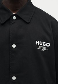 Μαύρο πουκάμισο με κουμπιά και γιακά, με λευκό λογότυπο "HUGO" και κείμενο "MCMXCIII Registered Trademark Since 1993" στο στήθος.