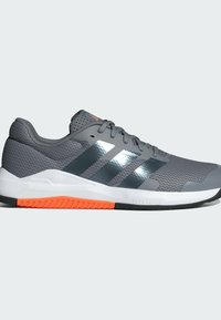 Zapatilla deportiva gris con parte superior texturizada, rayas metálicas plateadas, acento de suela anaranjada y una entresuela blanca acolchada.
