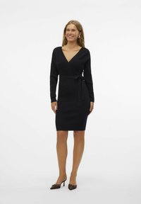 Abito avvolgente nero a coste con maniche lunghe, scollatura a V profonda e cintura con fiocco in vita. Il modello lo abbina a scarpe con tacco marroni.