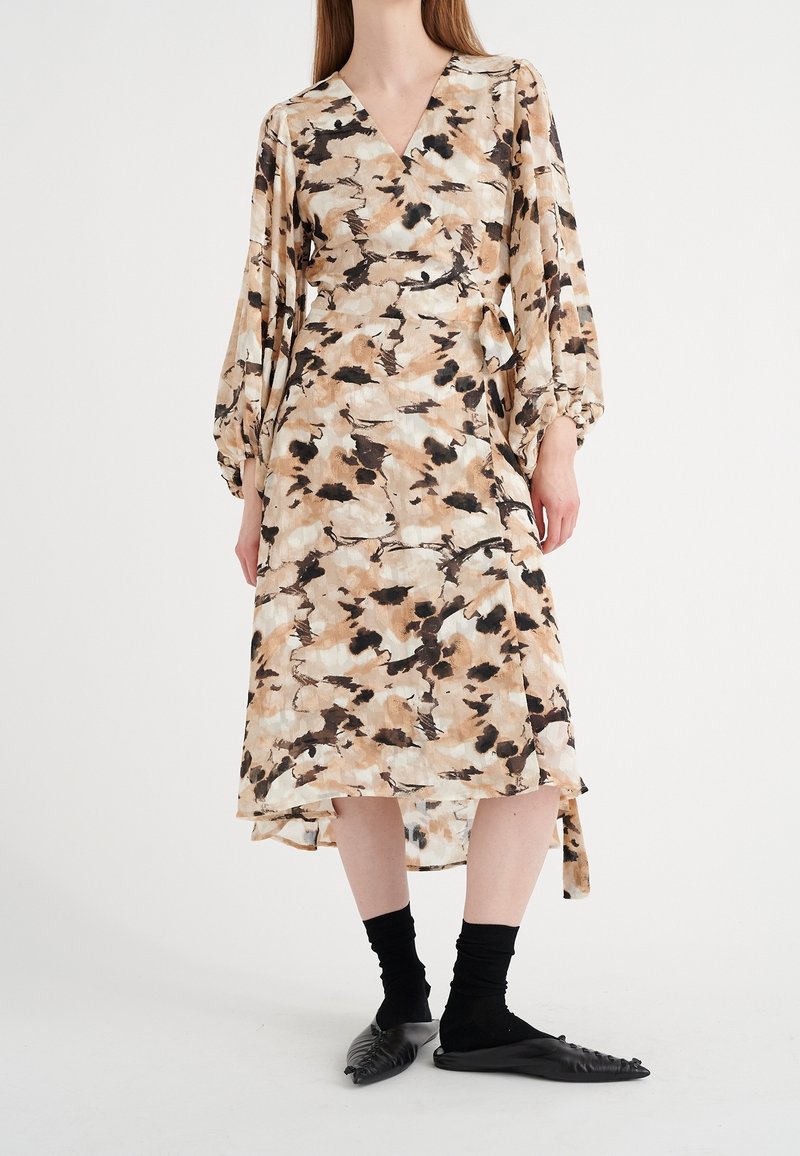 Femme portant une robe portefeuille midi à manches longues avec un imprimé abstrait beige, noir et blanc, associée à des chaussettes noires et des chaussures plates noires à bouts pointus.