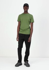 Green cotton t-shirt with short sleeves and a small logo on the chest, paired with black pants and gray sneakers. Black cap worn.
