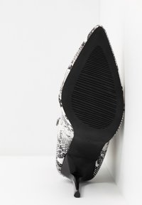 Chaussure à talon haut à bout pointu en cuir façon peau de serpent noir et blanc, vue de la semelle vers le haut sur fond blanc.