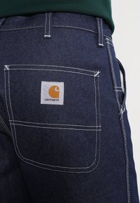 Dark denim jeans with white stitching, featuring a back pocket and a square tag with an orange logo and "Carhartt" text.