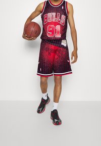 Maillot de basket-ball rouge et noir avec le texte "BULLS" et "91", présentant des rayures verticales. Shorts assortis avec un design éclaboussé, logo, et chaussures de sport noires.