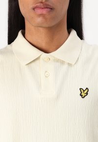 Polo-shirt crème à texture en jersey, avec un tissu côtelé, trois boutons et un logo d'aigle noir et jaune sur la poitrine gauche.