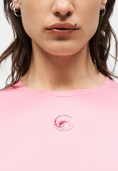 Κάτω μέρος γυναικείου προσώπου με γυαλιστερά χείλη, ελιά πάνω από το χείλος, φοράει κρίκους στα αυτιά και ροζ πουκάμισο με μικρό ροζ σχεδιασμό σπειροειδούς παγιέτας.