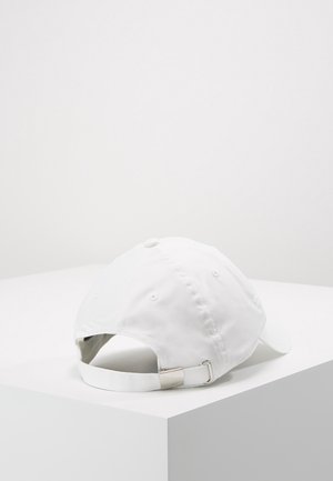 Casquette de baseball blanche avec sangle réglable à boucle métallique, placée sur une surface d'angle blanche sur fond blanc uni.