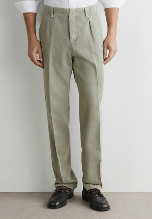 Mann trägt hellolivfarbene plissierte Hose mit schwarzen Lederschnürschuhen und weißem Langarmhemd vor einer einfarbigen Wand.