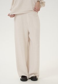 Light beige wide-leg trousers feature a textured fabric with subtle pattern detailing. The waistband is smooth, and the hem is loosely fitted over black shoes.