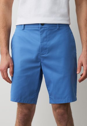 Mand iført ensfarvede blå shorts til knæene og en hvid t-shirt, stående med armene afslappet ned langs kroppen.