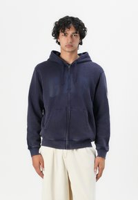 Sudadera con cremallera de color azul marino con un gran logo de "GAP" descolorido en el pecho. Tiene un bolsillo frontal y capucha ajustable con cordón. Tela suave.