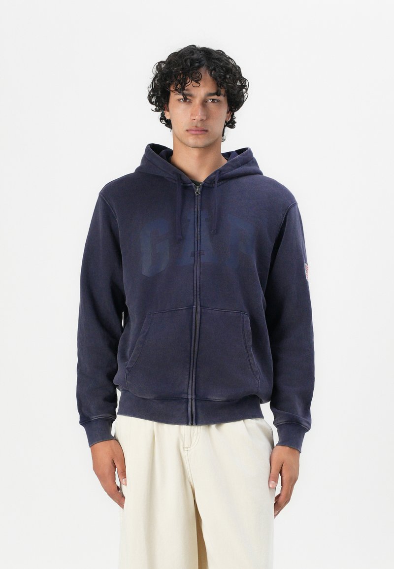 Sudadera con cremallera de color azul marino con un gran logo de "GAP" descolorido en el pecho. Tiene un bolsillo frontal y capucha ajustable con cordón. Tela suave.