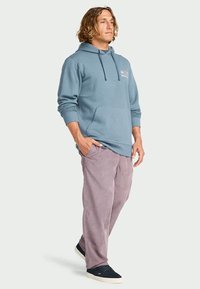 Homem com cabelo até aos ombros, vestido com um casaco azul, calças lilás e ténis pretos, em pé com as mãos relaxadas, olhando de lado.