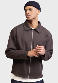 Chaqueta marrón con cremallera y acabado texturizado, diseño con cuello y un bolsillo frontal, llevada sobre una camisa blanca y jeans azules.