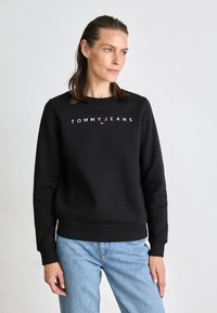 Felpa nera con logo bianco "TOMMY JEANS", colletto crew, polsini e orlo ribattuti, abbinata a jeans azzurri chiari, indossata in modo casual.