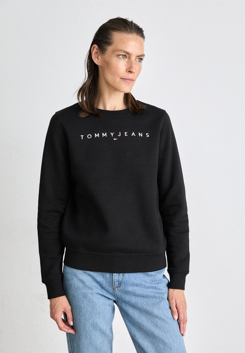 Felpa nera con logo bianco "TOMMY JEANS", colletto crew, polsini e orlo ribattuti, abbinata a jeans azzurri chiari, indossata in modo casual.