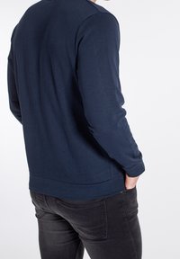 Marineblå langærmet skjorte med rund halsudskæring, blød tekstur og slank pasform. Båret over mørke jeans, med et sideblik der viser diskrete syninger.