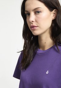 T-shirt di cotone viola con scollatura a girocollo e maniche corte, caratterizzato da un piccolo logo bianco nella zona sinistra del petto. Texture liscia.