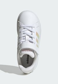 Bílý kožený teniska s texturovaným jazykem, bílými tkaničkami a třemi irizujícími proužky po straně. Uvnitř má měkkou, polstrovanou podšívku.