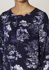 Blusa blu navy con un motivo floreale e boschivo in tonalità di viola e bianco; presenta un collo rotondo e un dettaglio arricciato nella parte frontale.