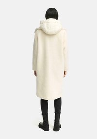 Lange, crèmekleurige jas met een pluche textuur en een fluffy, crèmekleurige capuchon. Voorzien van lange mouwen en een recht gesneden ontwerp. Zwarte leggings en laarzen dragen.
