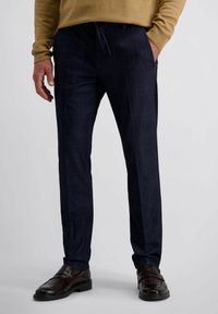Man in een donkerblauwe geruite broek, bruine leren instappers en een camel kleurige trui met handen in en naast de zakken.