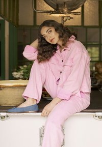 Roza denim jakna in hlače s sproščenim krojem. Modri ploski čevlji. Valoviti lasje, sedeča na pultu z rustikalno skledo v ozadju.