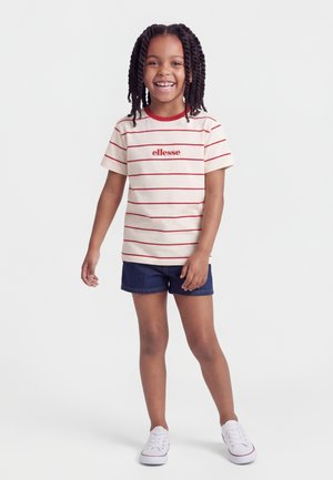 Χαμογελαστό παιδί με πλεξούδες, που φοράει κρεμ πουκάμισο με κόκκινες ρίγες και λογότυπο, σκούρο μπλε σορτς και λευκά αθλητικά παπούτσια, στέκεται σε απλό φόντο.