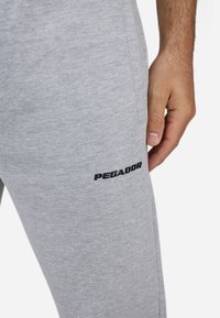 Harmaat verkkarit, jotka on valmistettu pehmeästä puuvillasekoituksesta. Vasemmassa reidessä on brodeerattu musta "PEGADOR" -logo, ja ne on muotoiltu kapenevaksi.