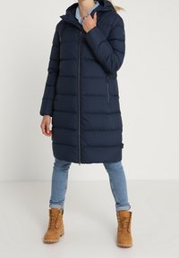 Mörkblå puffjacka med huva, horisontell sömnad, framsida med dragkedja och sidofickor. Bärs med ljusblå jeans och beige stövlar.