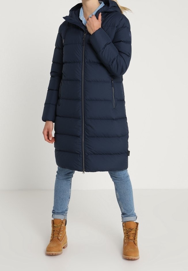 Mörkblå puffjacka med huva, horisontell sömnad, framsida med dragkedja och sidofickor. Bärs med ljusblå jeans och beige stövlar.