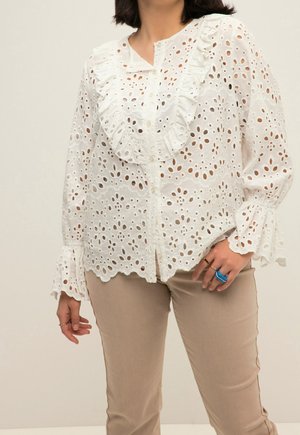 Femme portant un chemisier en dentelle blanche à œillets avec col et poignets volantés, associé à un pantalon beige et des bagues bleues et dorées à la main droite.
