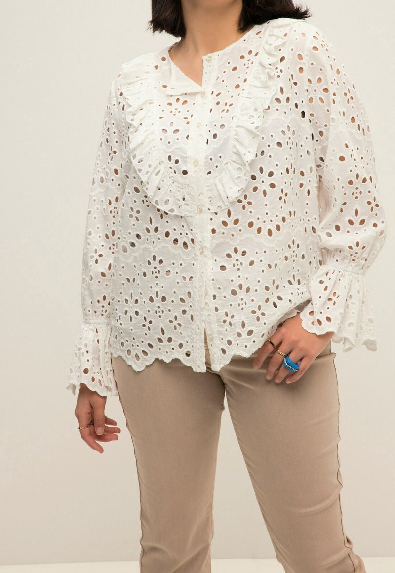 Femme portant un chemisier en dentelle blanche à œillets avec col et poignets volantés, associé à un pantalon beige et des bagues bleues et dorées à la main droite.