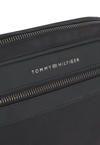 Μαύρη υφασμάτινη τσάντα με δύο μεγάλους μαύρους φερμουάρ και ασημί λογότυπο Tommy Hilfiger ανάμεσά τους.