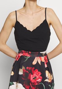 Svart scallop-formad linne med tunna axelremmar, parat med en blommig kjol med stora röda och gula blommor på en svart bakgrund.