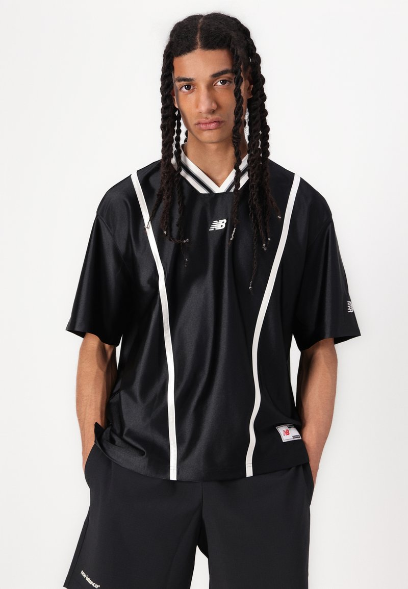 Maillot de sport noir en tissu brillant avec des rayures blanches sur les côtés, un col en V et un logo sur la poitrine. Porté avec un short noir.